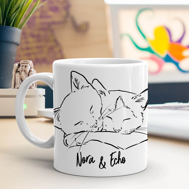 May include: White ceramic mug with a black line drawing of two cats sleeping on a pillow. The text "Nora & Echo" is printed below the cats.
