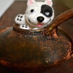 Op de afbeelding: Een witte en zwarte vilt hond met roze oren die uit een bruine keramische theepot kijkt.
