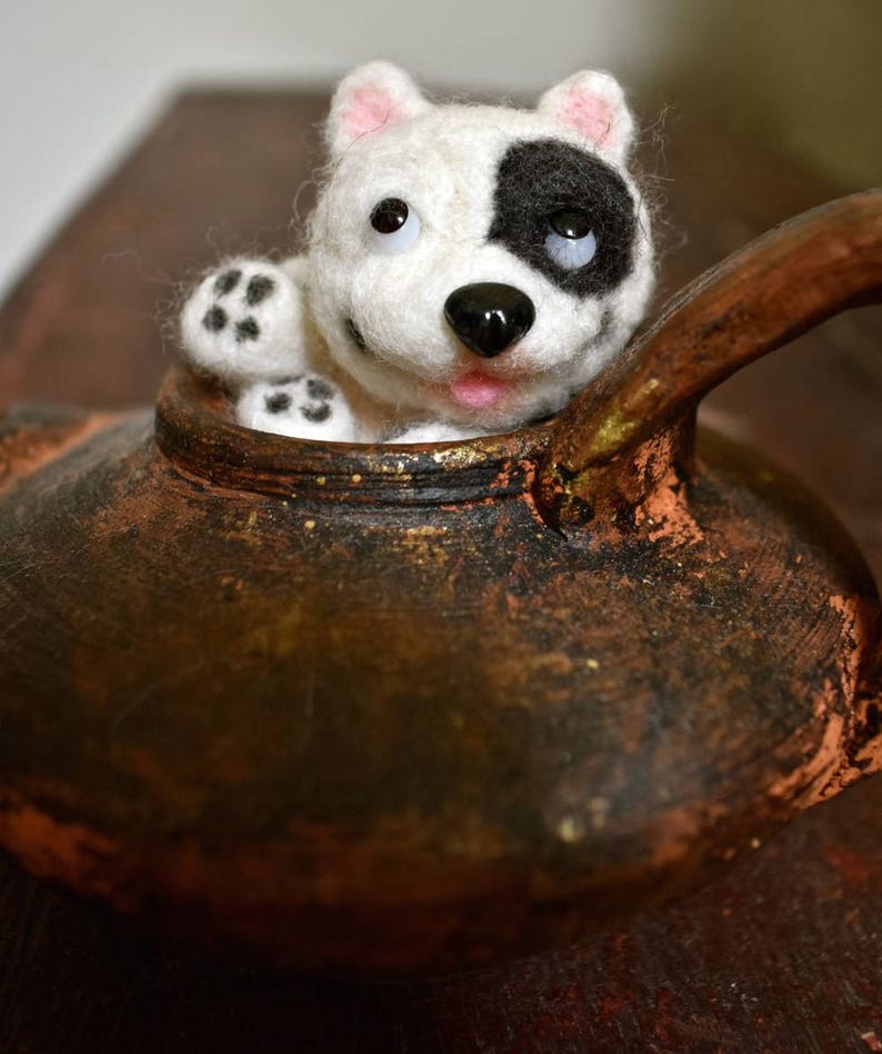 Op de afbeelding: Een witte en zwarte vilt hond met roze oren die uit een bruine keramische theepot kijkt.
