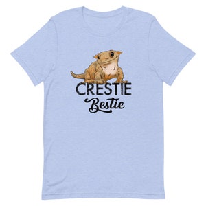 May include: Light blue t-shirt with a cartoon crested gecko and the text "Crestiebestie" in black.