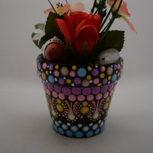 May include: A hand-painted flower pot with a black base and colorful dot patterns. The pot contains a red rose, other flowers, and decorative figurines. Green stems and leaves add to the arrangement. A charming decorative item.