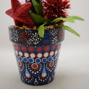 May include: A small terracotta pot decorated with red, white, and blue fireworks and geometric patterns. The pot is filled with faux greenery, a red glitter star, and a red pom-pom. The text "Option 1" is visible in the upper right corner of the image.