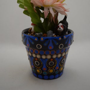 May include: A small, hand-painted terracotta pot with a black and blue geometric design. The pot is filled with small rocks and a small plastic dog figurine. The pot is decorated with yellow, pink, and white flowers.