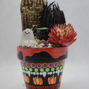 May include: A hand-painted Halloween-themed flower pot. The black pot features a decorative pattern of pumpkins, dots, and other spooky designs. It is filled with small stones, a ghost figurine, a small cauldron, a red succulent, and a tombstone that reads "RIP".
