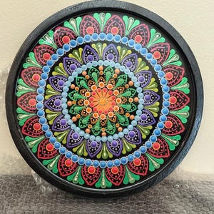 May include: A round, black-framed mandala art piece. The mandala features a symmetrical design with a black background and vibrant colors, including red, green, blue, purple, and orange. The intricate pattern is composed of dots and petal-like shapes.