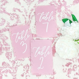 May include: Three pink table number cards with white calligraphy text. The cards are rectangular with a textured border. The numbers are 2, 3, and 7. White flowers are in the background.