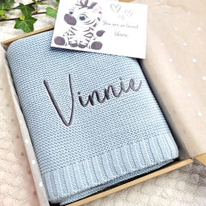 May include: A light blue knitted baby blanket with the name Vinnie embroidered in dark grey thread. The blanket is presented in a cardboard gift box that includes a white card featuring a cartoon zebra illustration and the text You are so loved Vinnie.