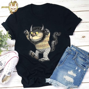 May include: Black t-shirt with a cartoon monster design. The monster has yellow eyes, horns, and a long tail. The shirt is paired with denim shorts and white trainers. The monster's torso is striped, and its legs are scale-like.