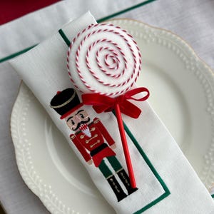 Op de afbeelding: Een witte servet met een groene rand is gevouwen in een driehoekige vorm en bedekt met een rood-witte snoepstok. Op de servet is een geborduurde notenkraker in rood en groen te zien. De servet ligt op een wit bord met een gouden rand.