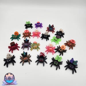 Peut inclure: Une collection de figurines d'araignées faites à la main. Chaque araignée est décorée de fleurs, de pierres précieuses et d'autres détails. Les araignées sont dans des tons de rose, de vert, de violet et de marron, avec des corps noirs.