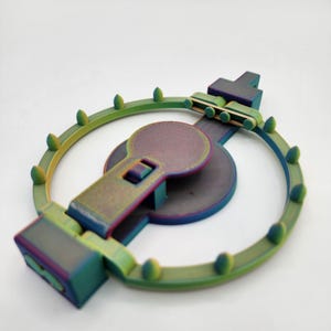 Puede incluir: Una trampa circular impresa en 3D, de color verde y morado iridiscente, con una palanca y una pequeña plataforma rectangular. La trampa tiene una serie de púas alrededor del perímetro.