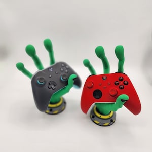May include: Two green 3D printed hands holding a black and a red video game controller. The hands are holding the controllers in a way that looks like they are giving a high five.