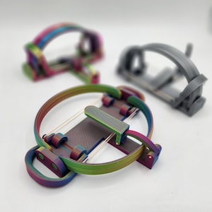 Può includere: Tre giocattoli fidget stampati in 3D. Il giocattolo in primo piano è color arcobaleno, con un design circolare con un centro rettangolare e una serie di archi di collegamento. Gli altri due giocattoli sono sfocati, uno arcobaleno e uno grigio.