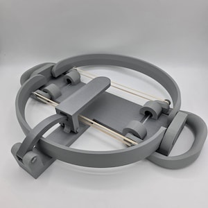May include: A gray 3D-printed object with a circular frame and a rectangular base. The design includes several curved and cylindrical components, connected by rubber bands. The object appears to be a mechanical or engineering model.