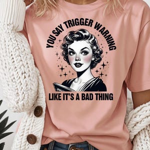 May include: A pink t-shirt with a black and white graphic of a woman with a retro hairstyle. The graphic says "You say trigger warning like it's a bad thing".