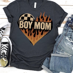 May include: Dark gray t-shirt with a heart-shaped design featuring a checkered flag pattern and flames. The words "BOY MOM" are printed in bold letters within the heart. Black and white sneakers and blue jeans are also visible.