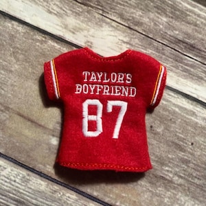 May include: A small, red jersey with white embroidered text that reads "TAYLOR'S BOYFRIEND" and the number "87". The jersey has white and gold stripes on the sleeves and neckline. The jersey is made of a felt-like material.