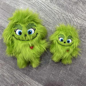 May include: Two plush green Grinch-like figures with shaggy fur. The larger figure has a smiling face with blue eyes and a red heart. The smaller figure has a similar design. The figures are standing upright.