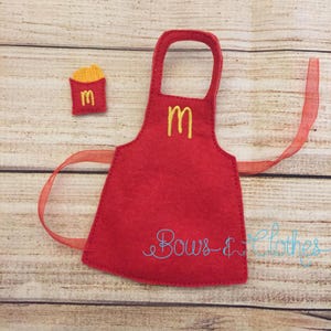 Puede incluir: Un delantal de fieltro rojo con un logotipo "M" amarillo y una bolsa de patatas fritas a juego. El delantal tiene una cinta y el texto "Bows & Clothes" en escritura azul. La bolsa de patatas fritas tiene un gráfico de patatas fritas amarillas.