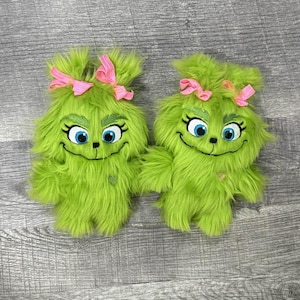 May include: Two plush green character dolls with pink bows, blue eyes, and smiling faces. The dolls have a furry texture and are designed to resemble a popular holiday character. The dolls are laying on a grey wooden surface.