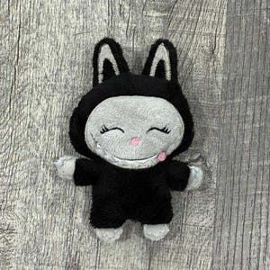 May include: A plush toy in the shape of a smiling cat wearing a black bunny suit. The cat's face is gray with black stitched eyes and a pink nose. The bunny suit has white accents on the ears and paws.