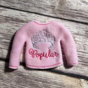 May include: A light pink felt sweater with long sleeves. The sweater features an embroidered silver crown design above the word "Popular" in a cursive font. The stitching is in a contrasting pink color.