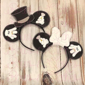 Op de afbeelding: Een paar zwarte en witte Mickey Mouse oren met een hoge hoed en een strik. De oren hebben een wit handschoendesign met een diamanten ring aan elke vinger.