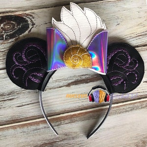 Op de afbeelding: Een zwarte haarband met muizenoren. De oren zijn zwart met paarse geborduurde tentakels. Een holografische strik met een gouden zeeschelp en witte veermotieven zit bovenop de haarband. Een kleine bedel van een koptelefoon is bevestigd.