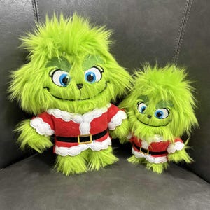 May include: Two plush Grinch dolls. The larger doll has green fur, blue eyes, and a red and white Santa-style outfit with a black belt. The smaller doll mirrors the larger one, sharing the same color scheme and design.