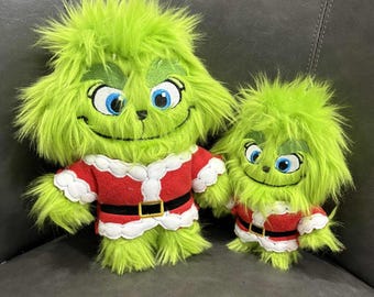 May include: Two plush Grinch dolls. The larger doll has green fur, blue eyes, and a red and white Santa-style outfit with a black belt. The smaller doll mirrors the larger one, sharing the same colour scheme and design.