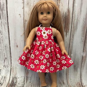 May include: A doll in a red dress patterned with white daisies. The dress features a white strap and an apron-style top. The doll has long blonde hair and is set against a wooden backdrop.