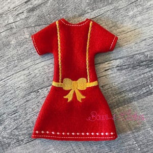 Peut inclure: Une robe rouge en feutre avec des manches courtes et un détail de nœud jaune. La robe a des bretelles brodées jaunes et un ourlet décoratif. La robe est de taille miniature, probablement pour une poupée ou un projet d'artisanat.