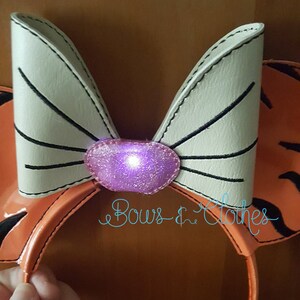 May include: A pair of orange and white mouse ears with a white bow and a purple light-up centre. The ears are made of faux leather and have a black stitched outline. The bow has a black stitched outline and a purple glitter centre. The text "Bows & Clothes" is visible in the lower right corner of the image.