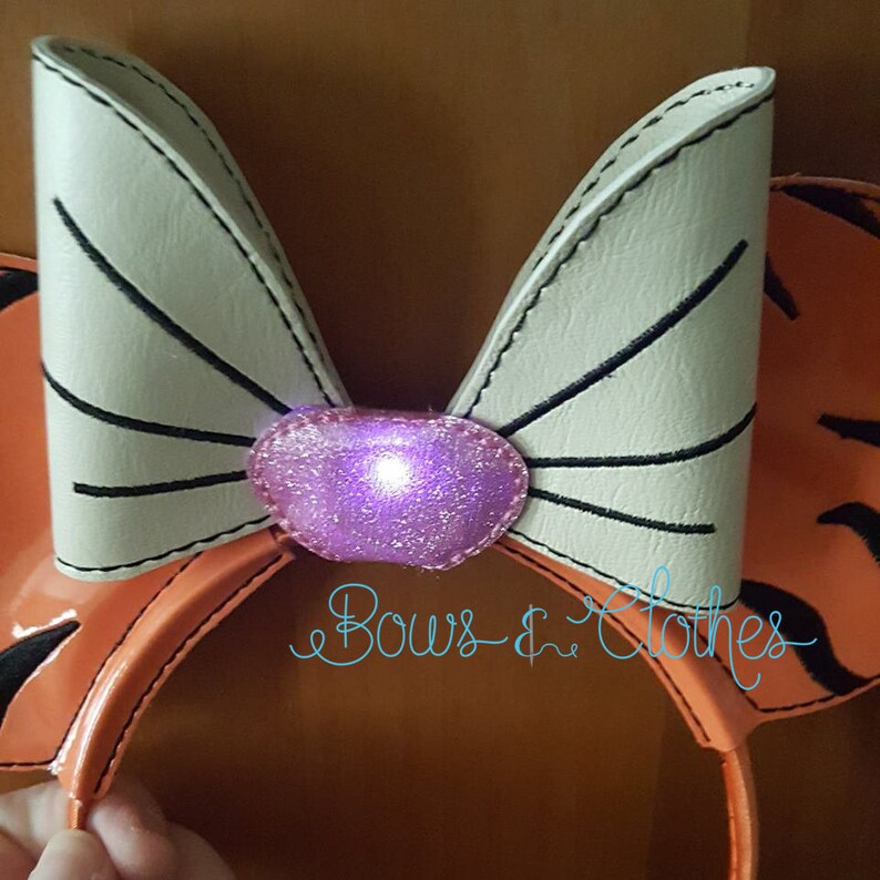 May include: A pair of orange and white mouse ears with a white bow and a purple light-up centre. The ears are made of faux leather and have a black stitched outline. The bow has a black stitched outline and a purple glitter centre. The text "Bows & Clothes" is visible in the lower right corner of the image.