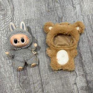 May include: Two plush toys: a grey monster with bunny ears and a brown bear costume. The monster has a large, smiling face with black eyes. The bear costume has a white belly and a hood. Both toys are on a wooden surface.