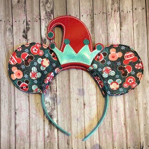 May include: Christmas themed Minnie Mouse ears headband with a red and green elf hat and a teal crown. The ears are covered in a green fabric with a pattern of candy canes, snowflakes, and Mickey Mouse heads.