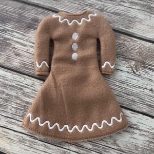 May include: A brown felt dress with long sleeves and a flared skirt. The dress is decorated with white, wavy stitching along the neckline, cuffs, and hem. Three white buttons are sewn down the front of the dress.