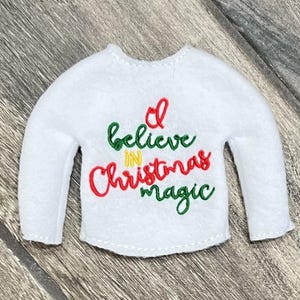 Peut inclure: Ornement de Noël en feutre blanc en forme de pull avec des manches longues. Le texte "I believe in Christmas magic" est brodé en fil rouge, vert et jaune. L'ornement est conçu pour ressembler à un mini pull.
