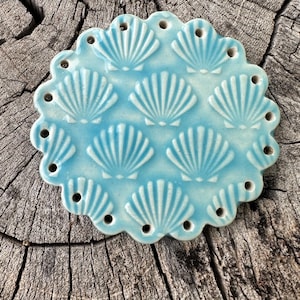 May include: A light blue ceramic coaster with a scalloped edge and a repeating pattern of seashells. The coaster has small holes around the edge.