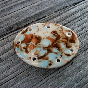 May include: Oval ceramic soap dish with drainage holes. The dish has a cream base with brown, orange, and light blue swirling patterns. The soap dish is designed to hold a bar of soap.