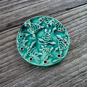 May include: A teal ceramic dish with a circular design featuring multiple sea turtle silhouettes. The dish has several small holes around the edge.