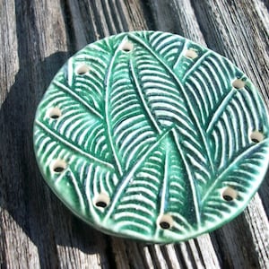 May include: A round, ceramic soap dish with a leaf design in shades of green and white. The dish has a series of small holes around the edge. The dish is placed on a weathered wooden surface.