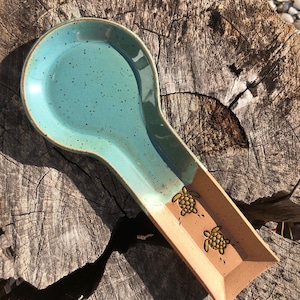 Peut inclure: Un repose-cuillère en céramique avec une surface bleu turquoise et un fini moucheté. Le manche est de couleur marron clair avec deux motifs de tortues. Le repose-cuillère est posé sur une surface en bois.