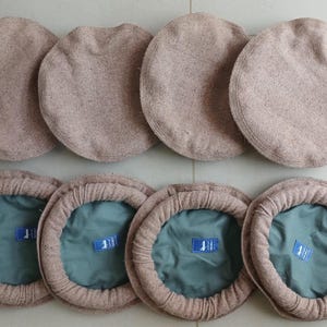Op de afbeelding: Een verzameling van acht beige Pakol-hoeden, een traditionele Afghaanse hoed. De hoeden zijn gerangschikt in twee rijen, met de bovenkant van de hoeden in de bovenste rij en de binnenkant van de hoeden in de onderste rij. De binnenkant van de hoeden is teal.