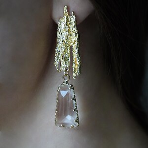 May include: Gold tone earrings with a textured, abstract design and a clear, faceted crystal drop.