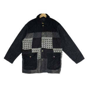 May include: A black and gray patchwork jacket with a collared neckline and button-up closure. The jacket features two front pockets and a mix of textured fabrics, including a geometric pattern. The jacket is displayed on a wooden hanger.