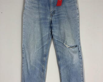 May include: Light blue denim jeans with distressed details, including a ripped knee and frayed pocket edges. The jeans hang from a wooden hanger, with a red tag attached. The jeans have a classic five-pocket design.