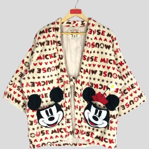 May include: A cream-colored Mickey Mouse themed robe with red and brown lettering. The robe has a tie closure and features two large embroidered Mickey Mouse faces on the front.