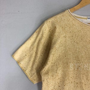 May include: A short-sleeved, crew neck t-shirt in a speckled tan color. The shirt has a white interior collar and the word "STONE" is visible on the front. The shirt is hanging on a wooden hanger against a gray background.