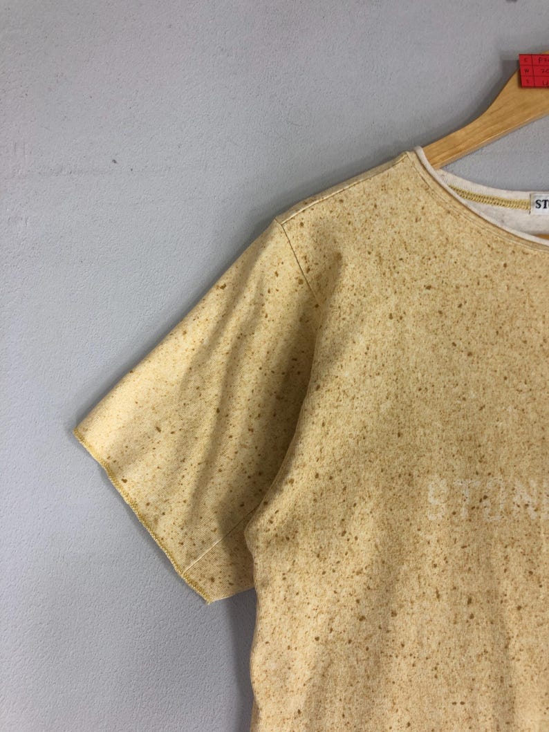 May include: A short-sleeved, crew neck t-shirt in a speckled tan color. The shirt has a white interior collar and the word "STONE" is visible on the front. The shirt is hanging on a wooden hanger against a gray background.